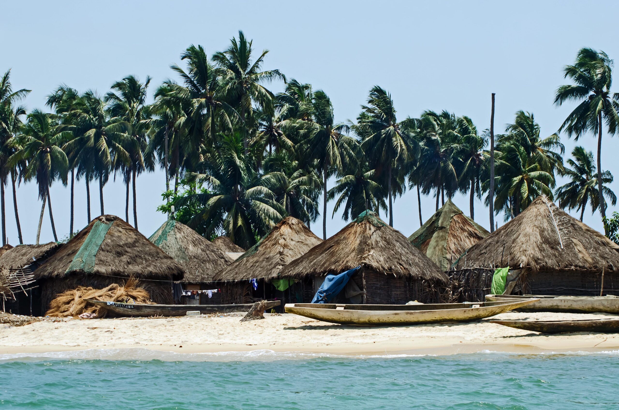 Thatched buildings and boats line a sandy waterfront beneath palm trees.