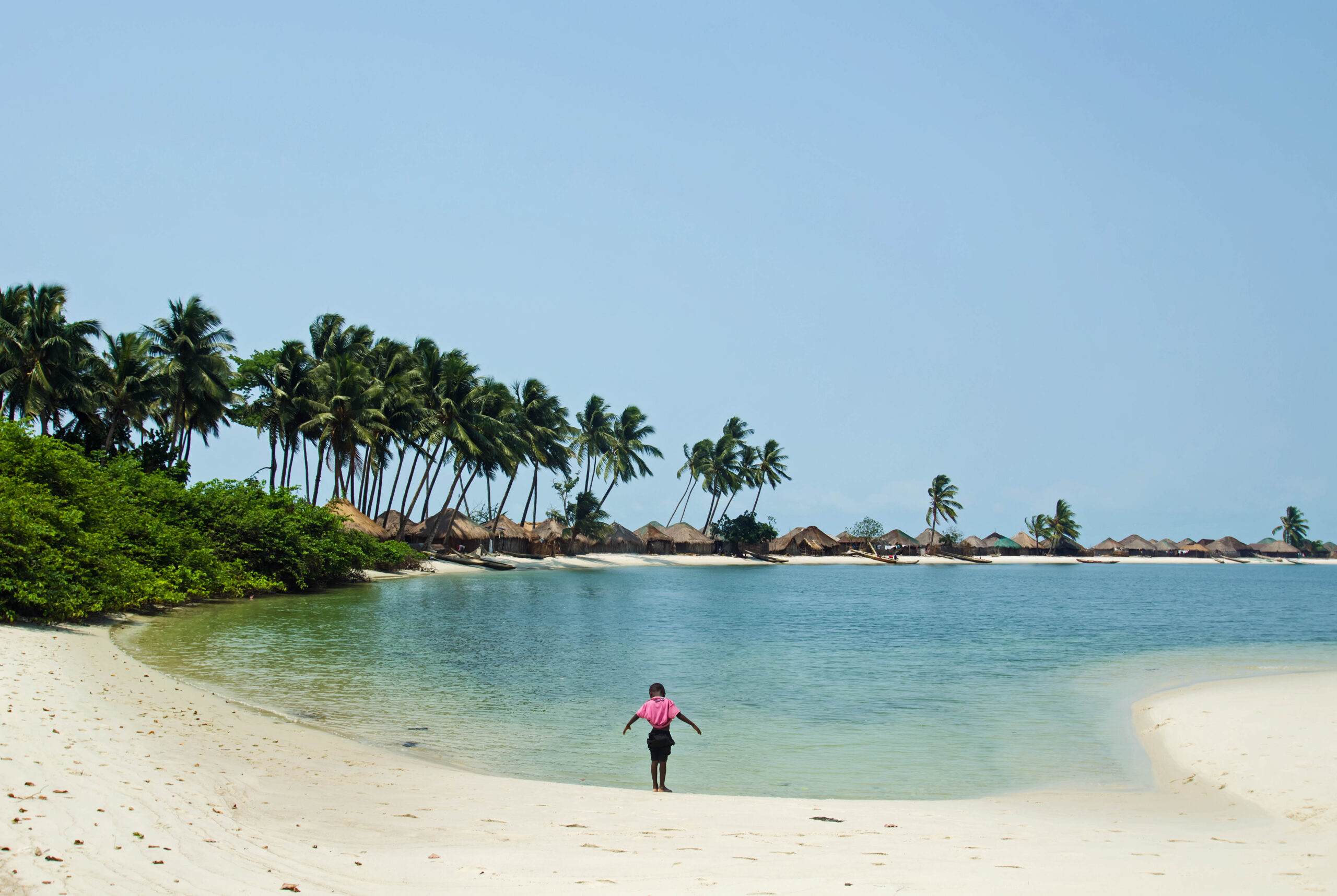 Person at the edge of a sandy bay with clear blue water, palm trees and distant huts.