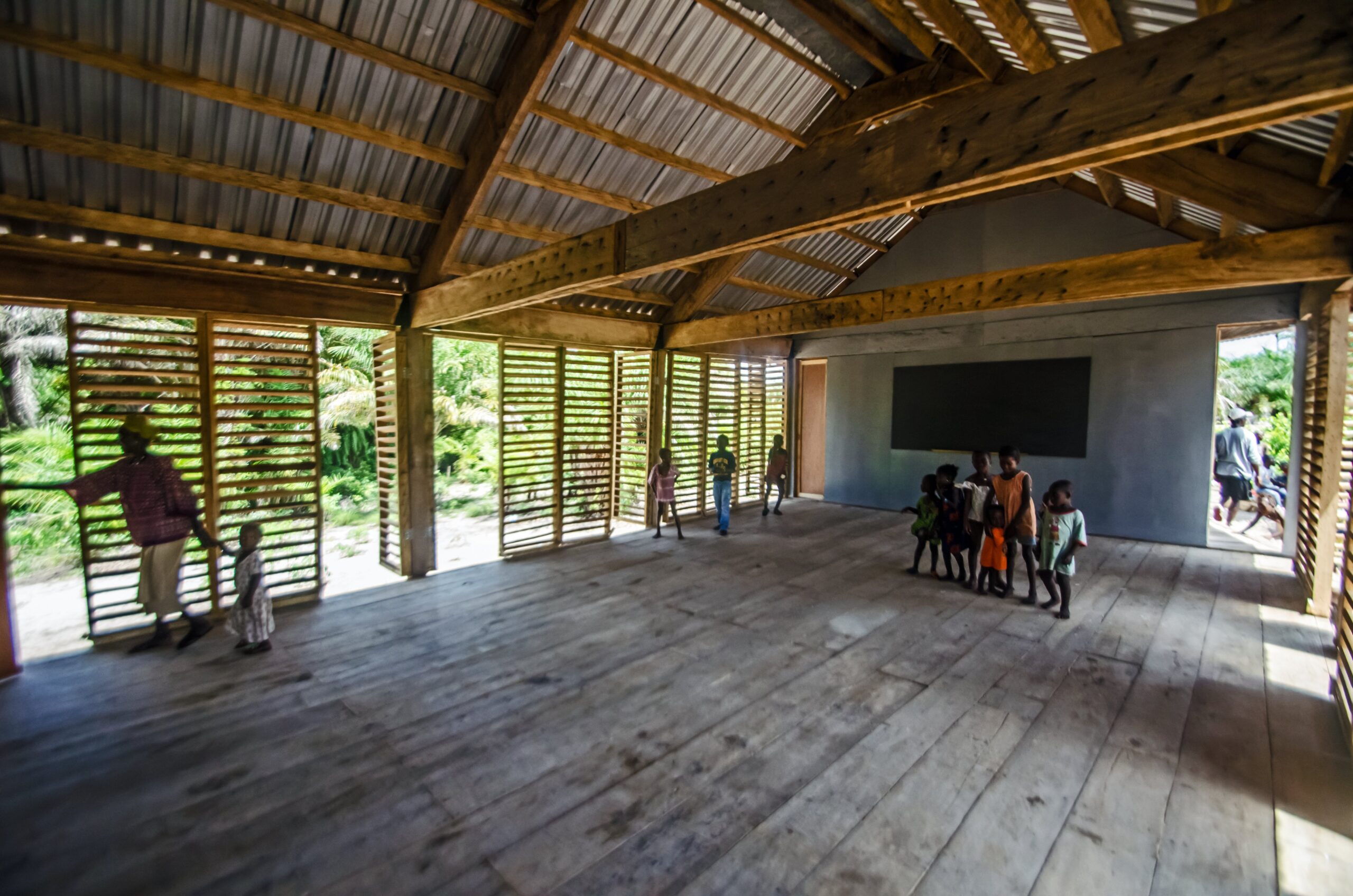 Wide classroom interior with timber floors, exposed roof framing, slatted walls and children near a chalkboard.
