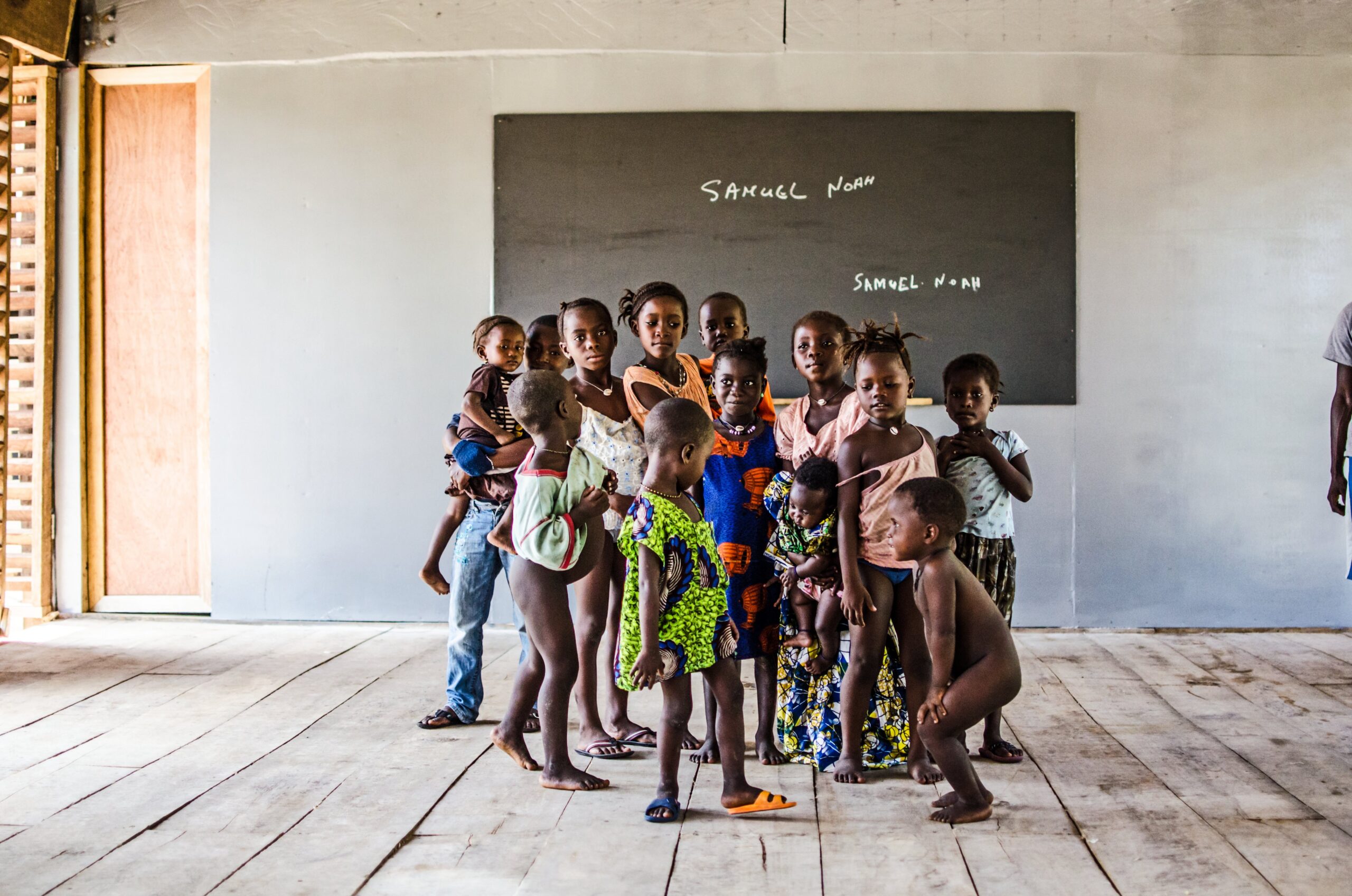 Group of children standing on a timber classroom floor in front of a chalkboard.