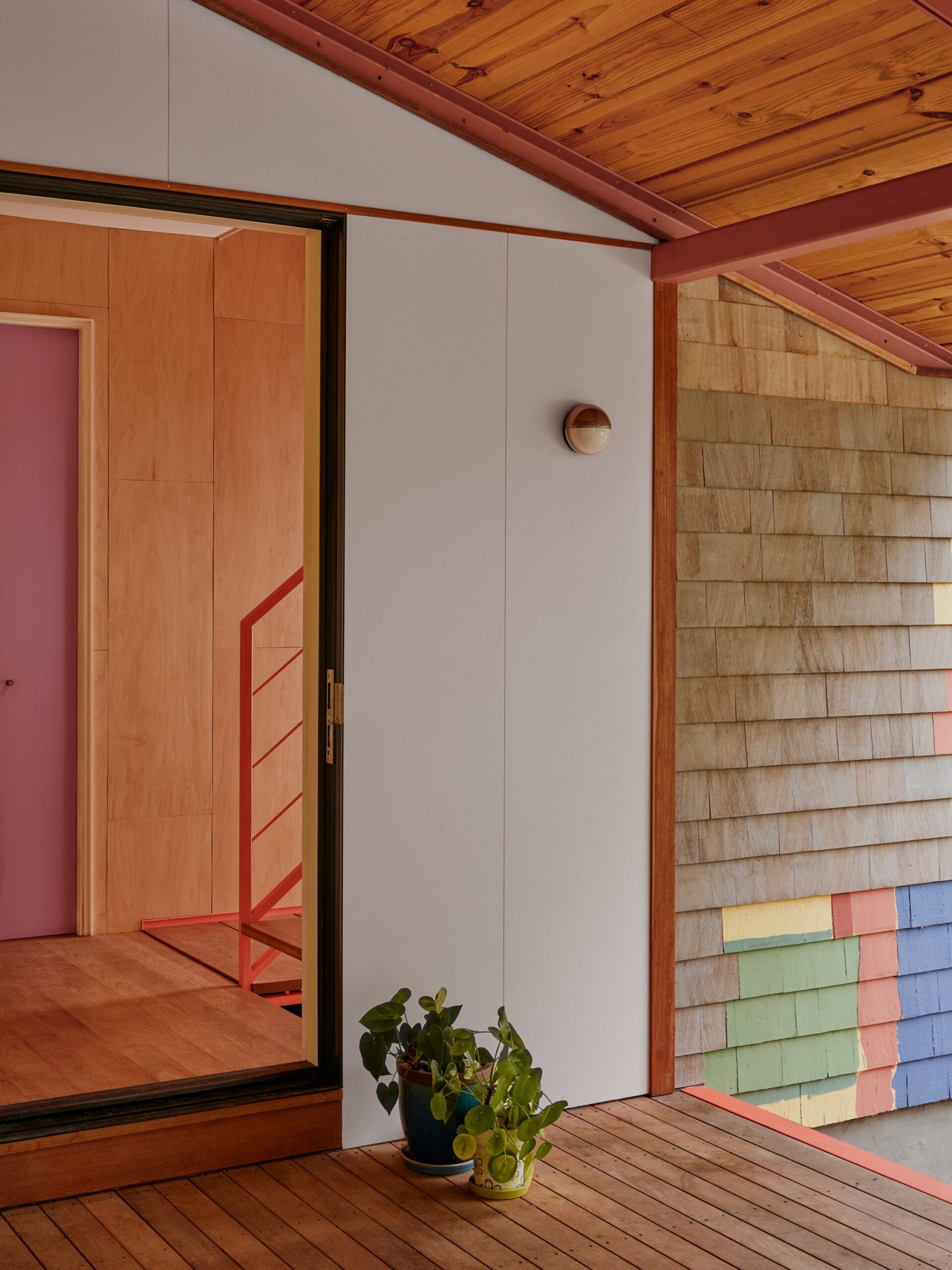 Wood-lined interior threshold with a red stair rail opens toward a shingled wall with colored panels.