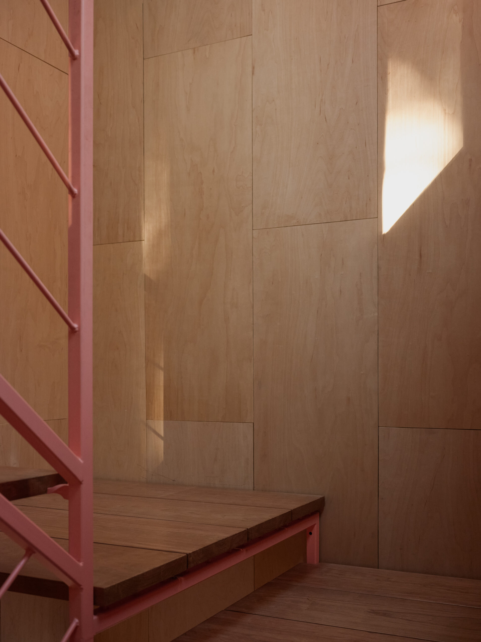 Wood-lined stair landing with a red metal railing and sunlight across the wall.