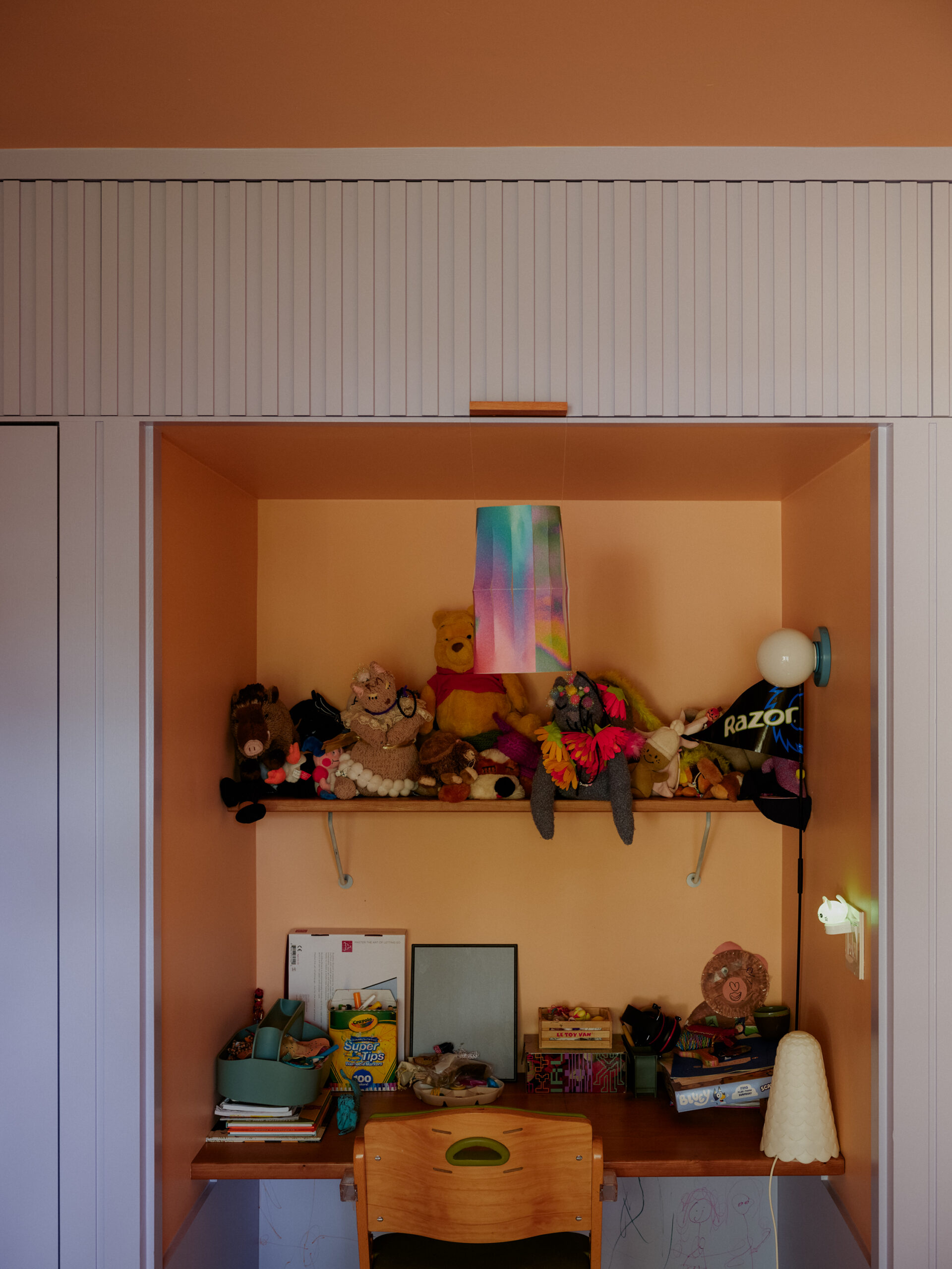 Orange recessed desk nook with shelves, books and toys framed by pale wall paneling.