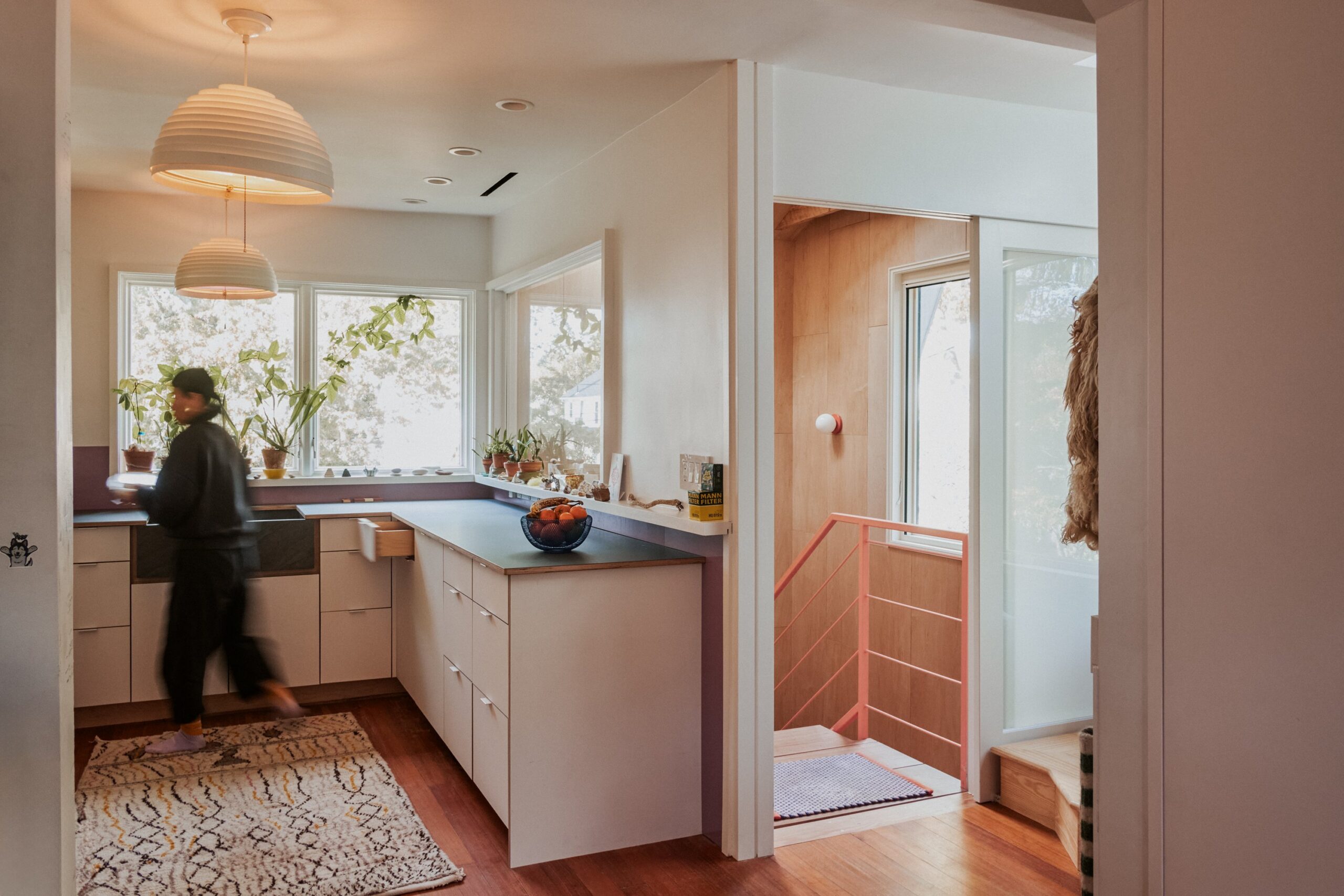 Kitchen with pale cabinets and pendant lights beside a wood-lined stair opening with a red rail.