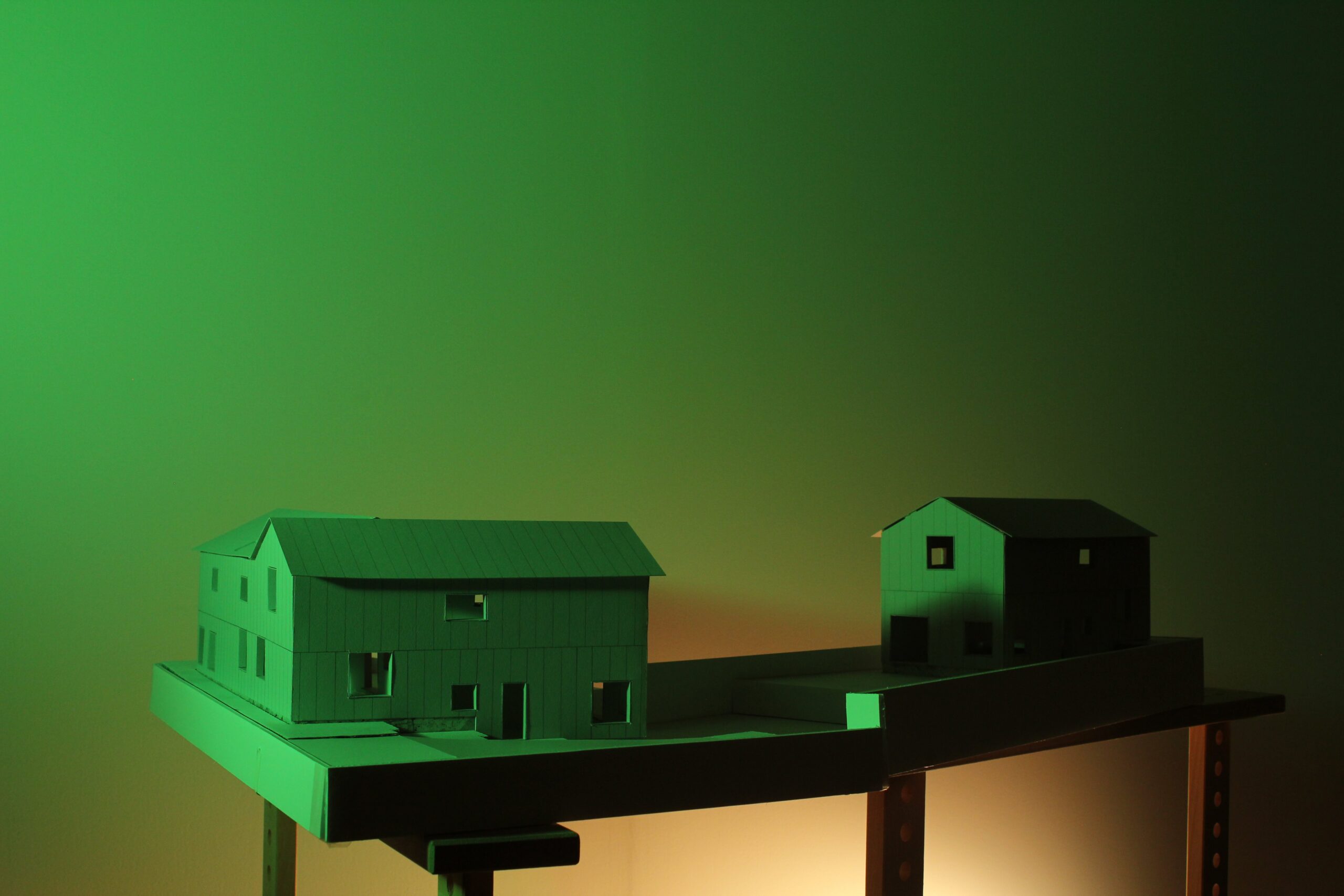 Two pitched-roof building models sit on an elongated base under green and warm lighting.