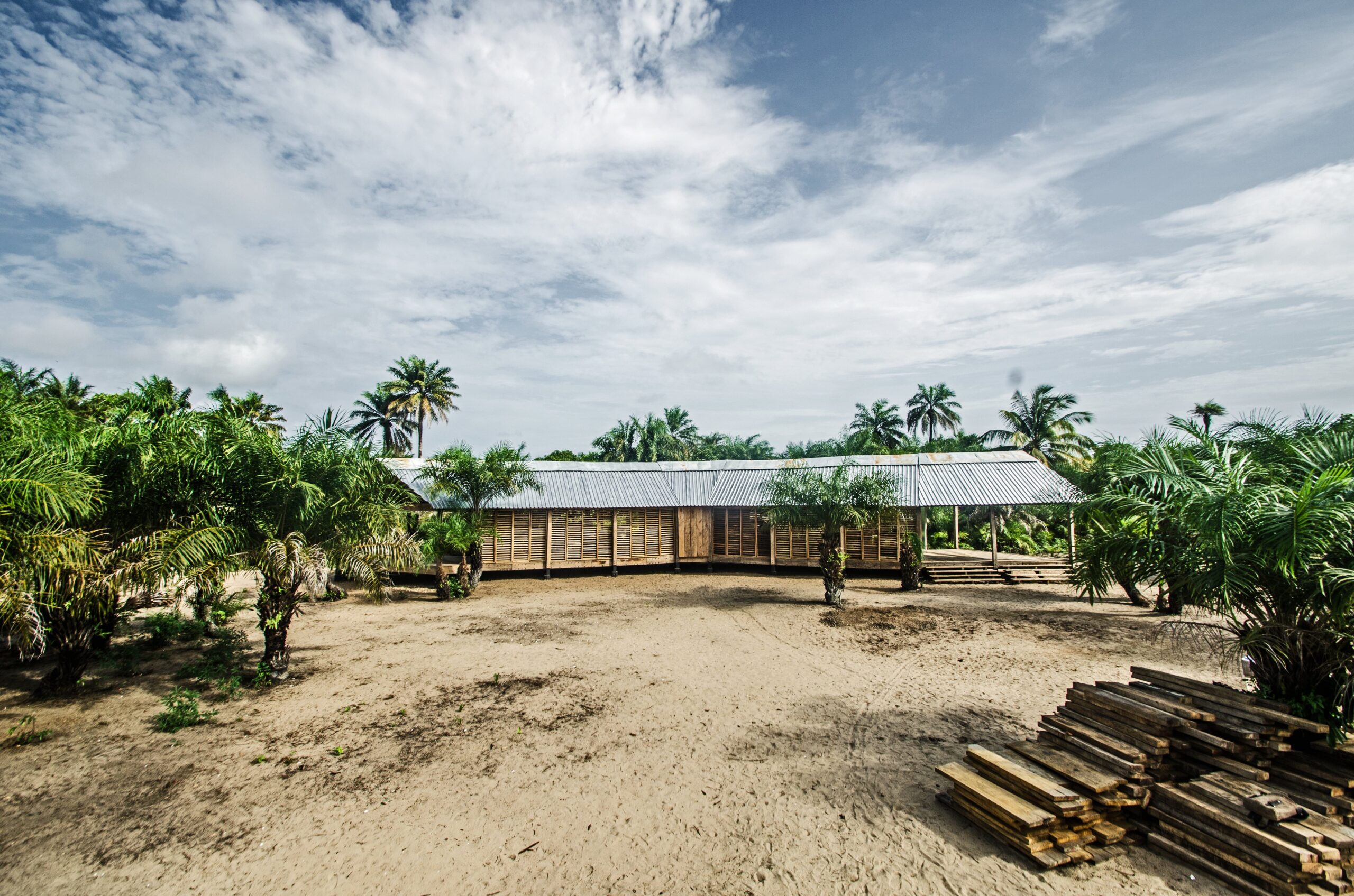 Long low school building with a pitched roof and slatted walls beside sandy grounds and palms.