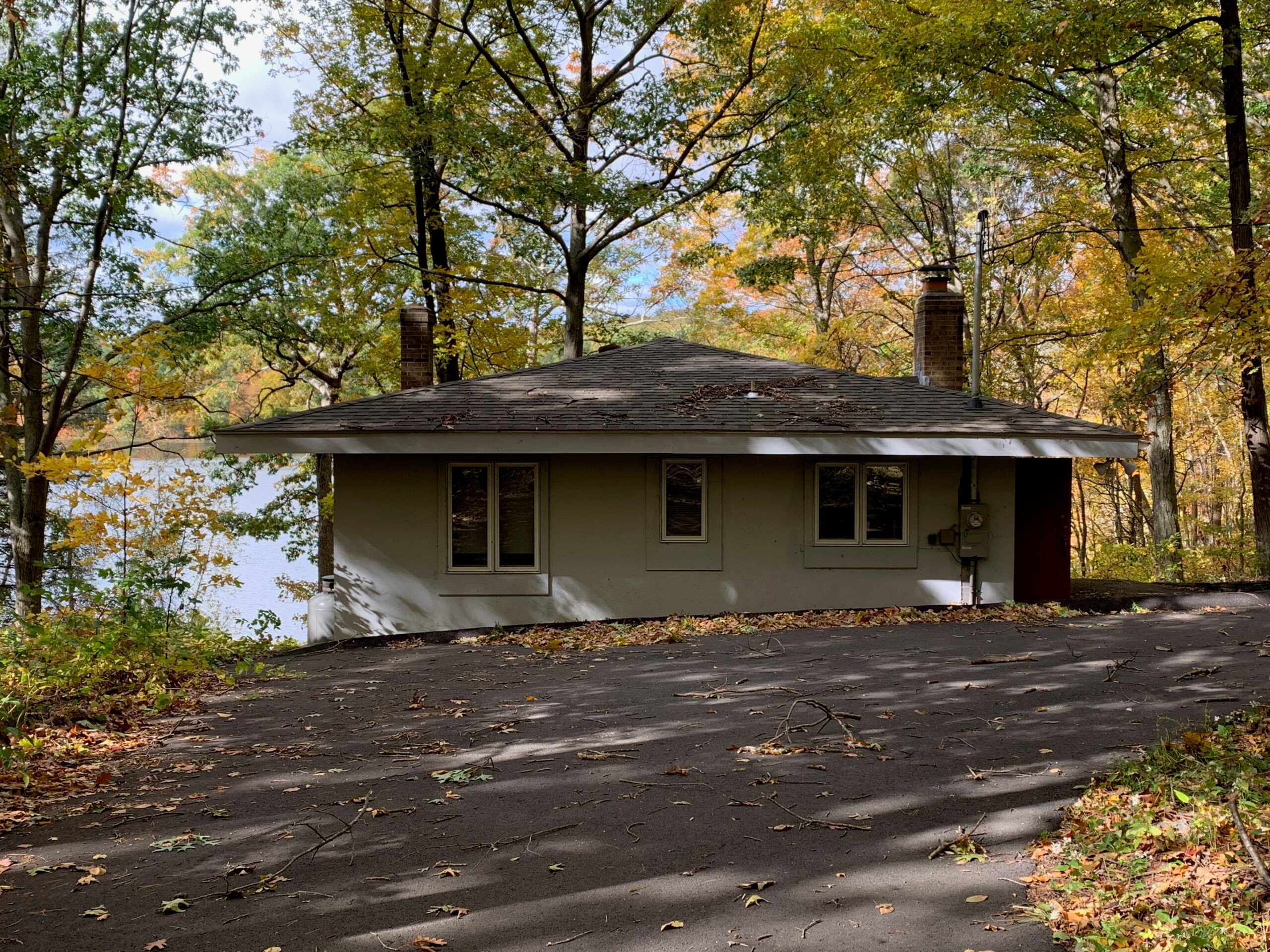 A low house with a hipped roof stands among autumn trees beside a paved drive.