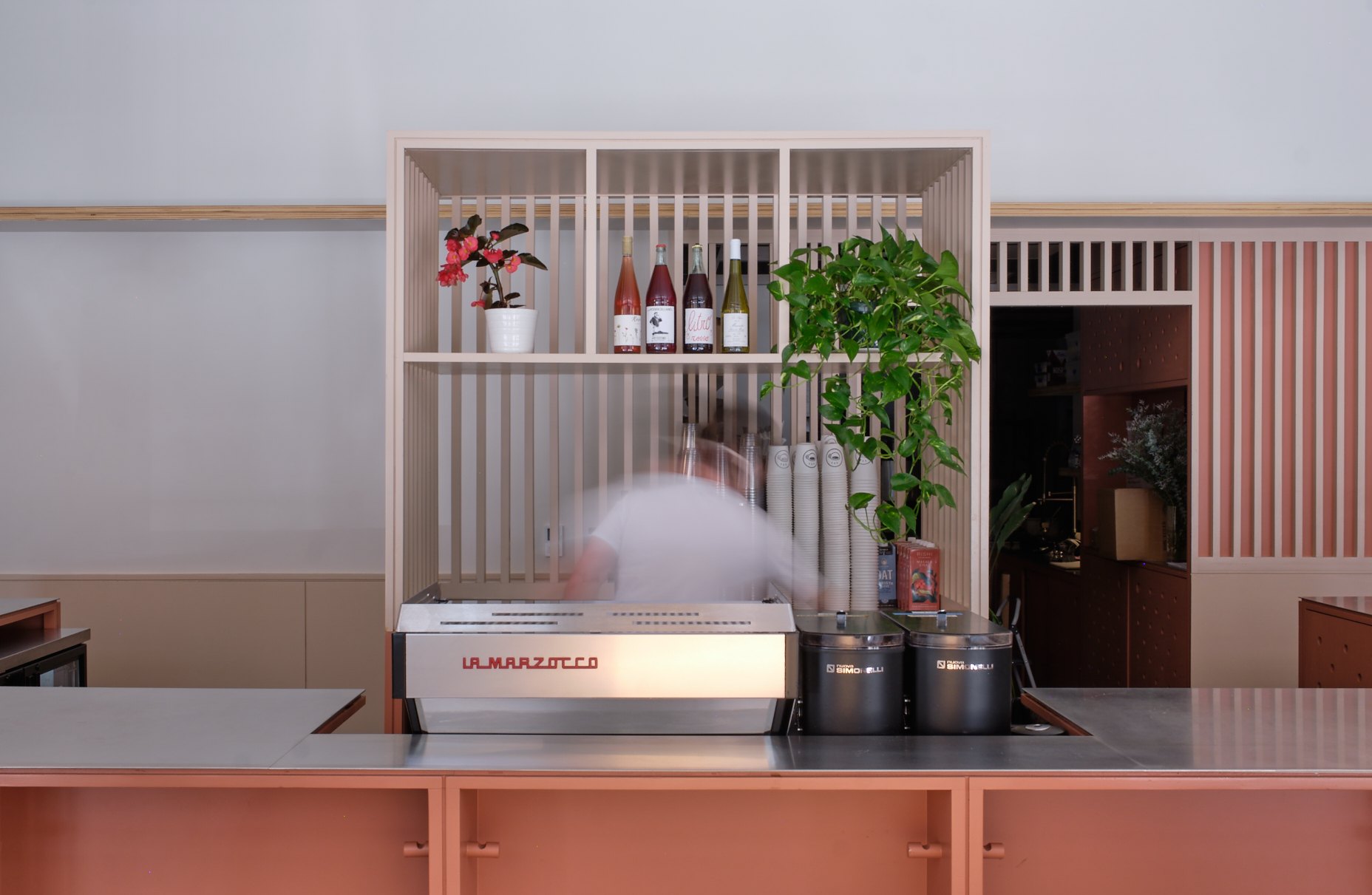 An espresso machine sits on a coral café counter in front of a slatted display with bottles and plants.
