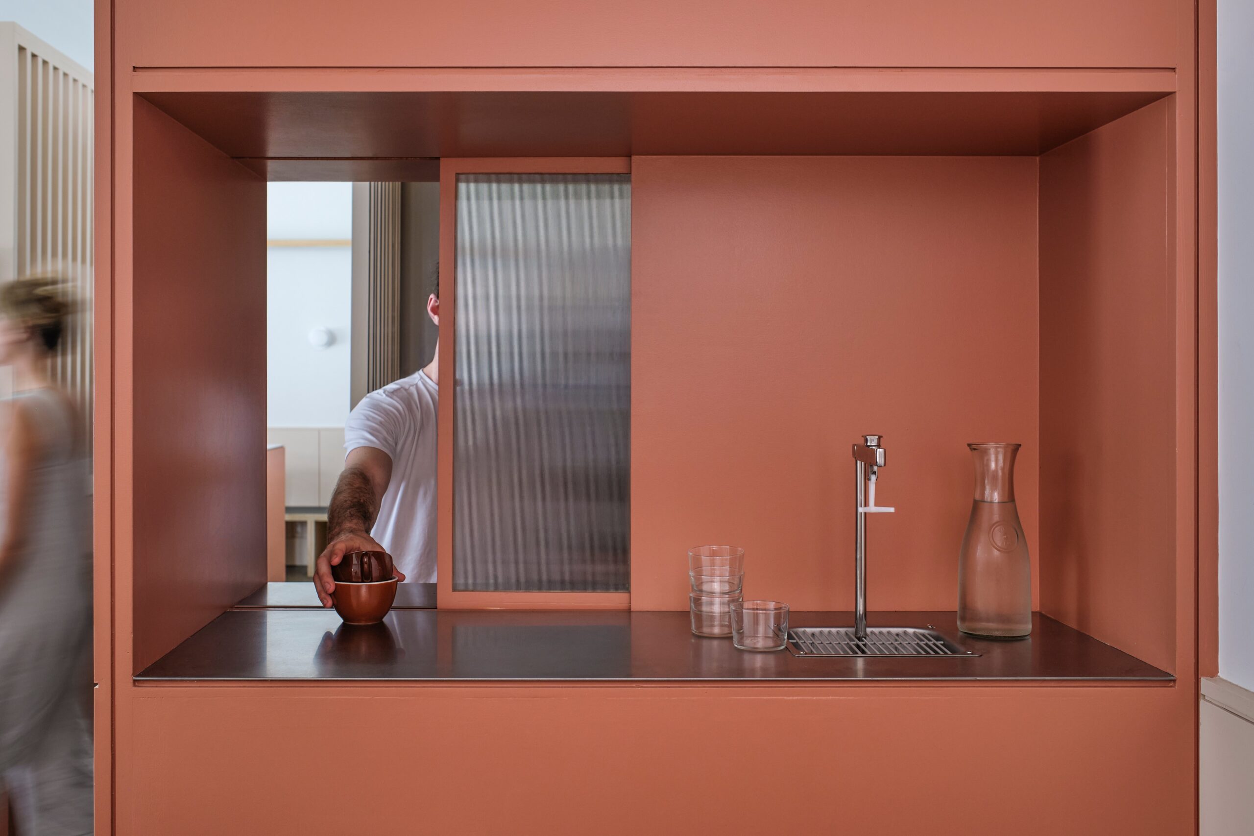 A recessed service station with a metal counter, tap and glassware is framed by coral cabinetry.
