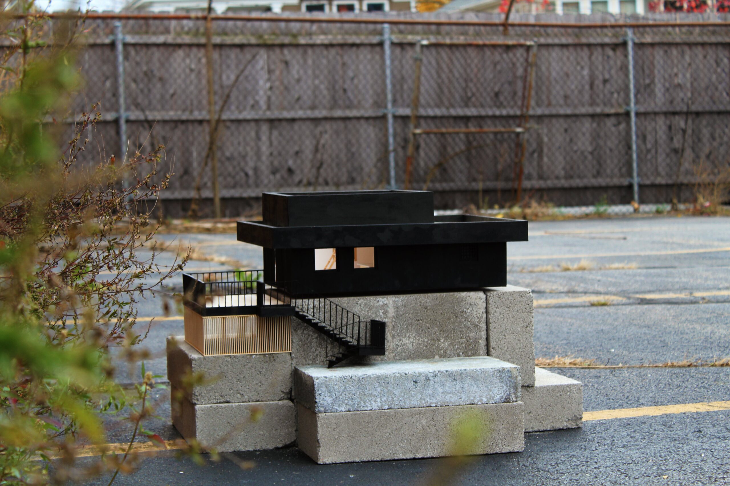Black house model and miniature deck railings sit on concrete blocks in an outdoor setting.