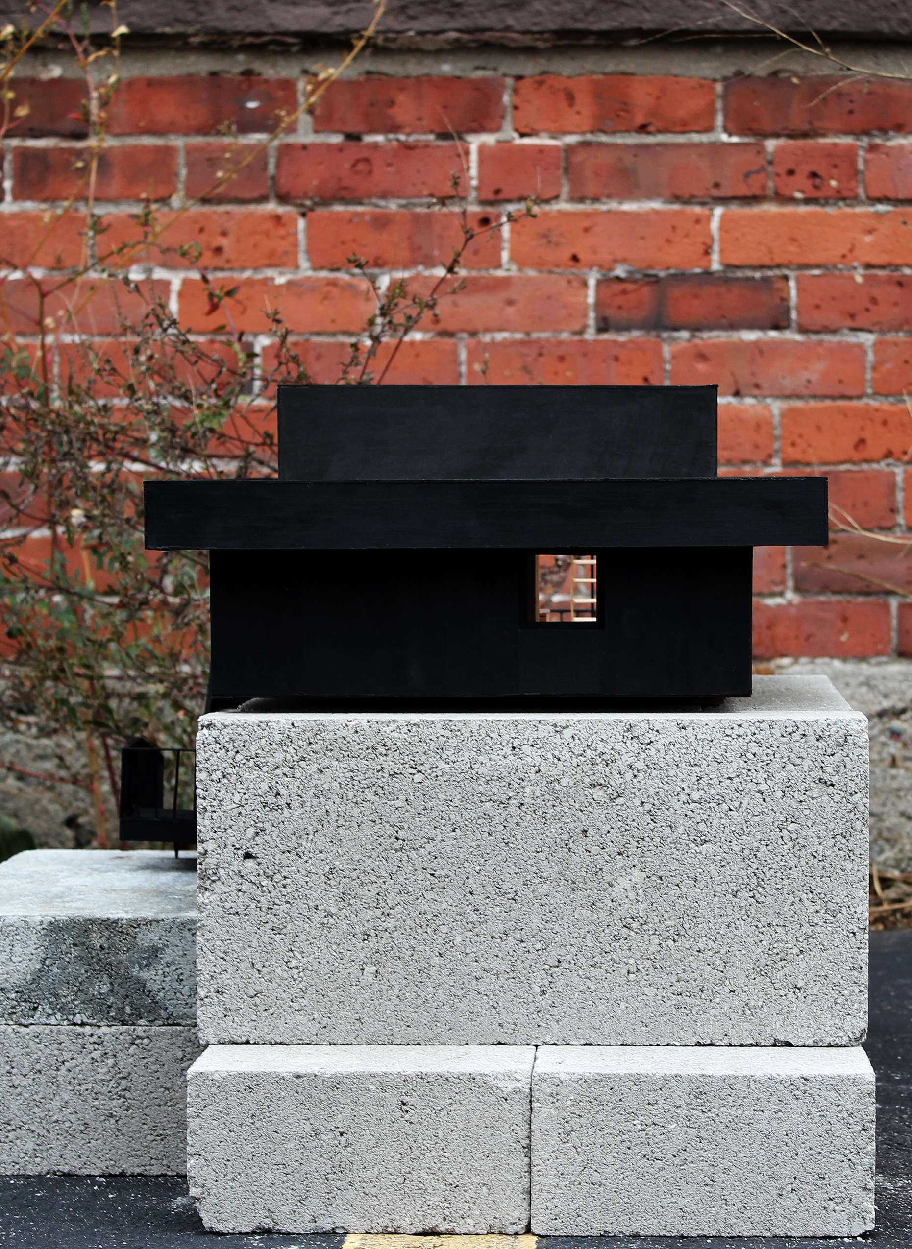 Black architectural model with a projecting horizontal roof rests on concrete blocks against a brick wall.