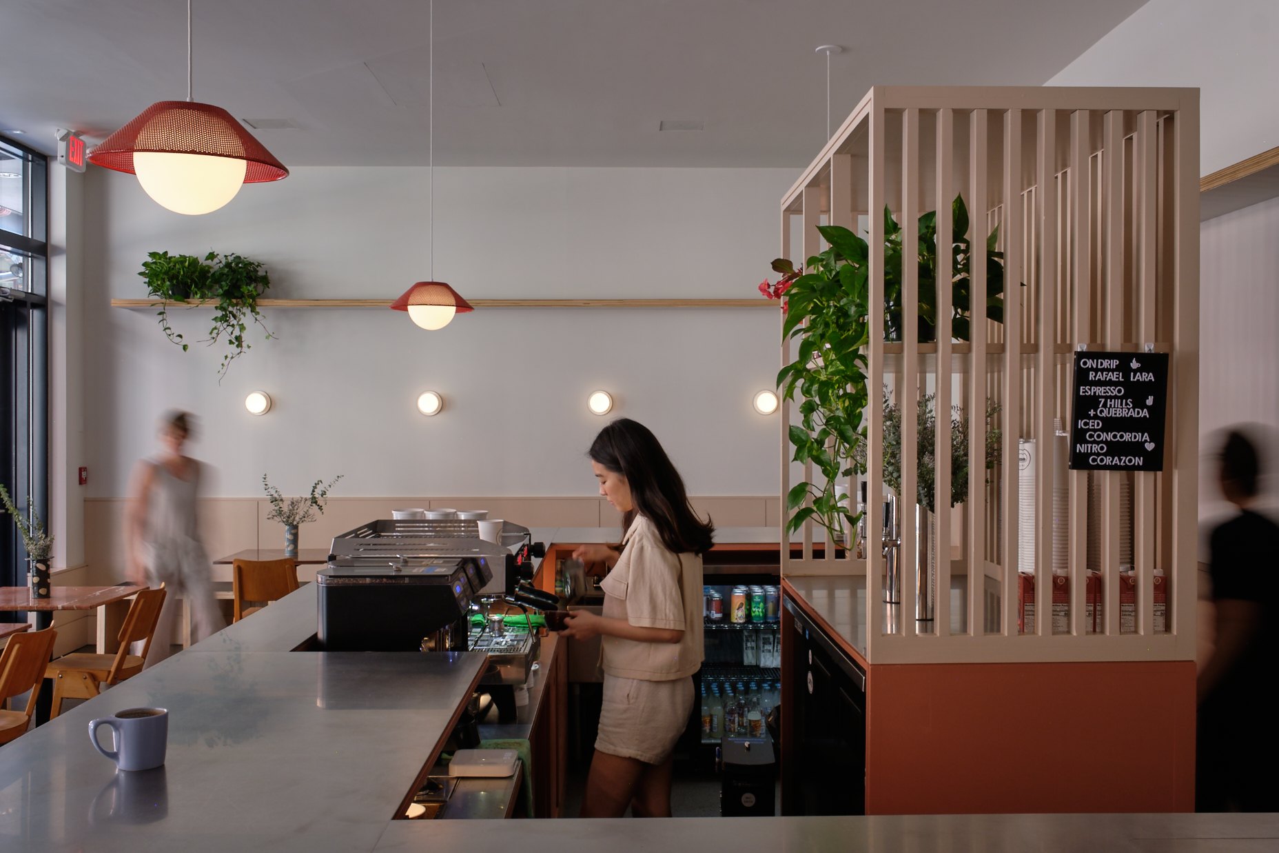 A café worker stands behind a counter beside a slatted partition, hanging plants and pendant lights.