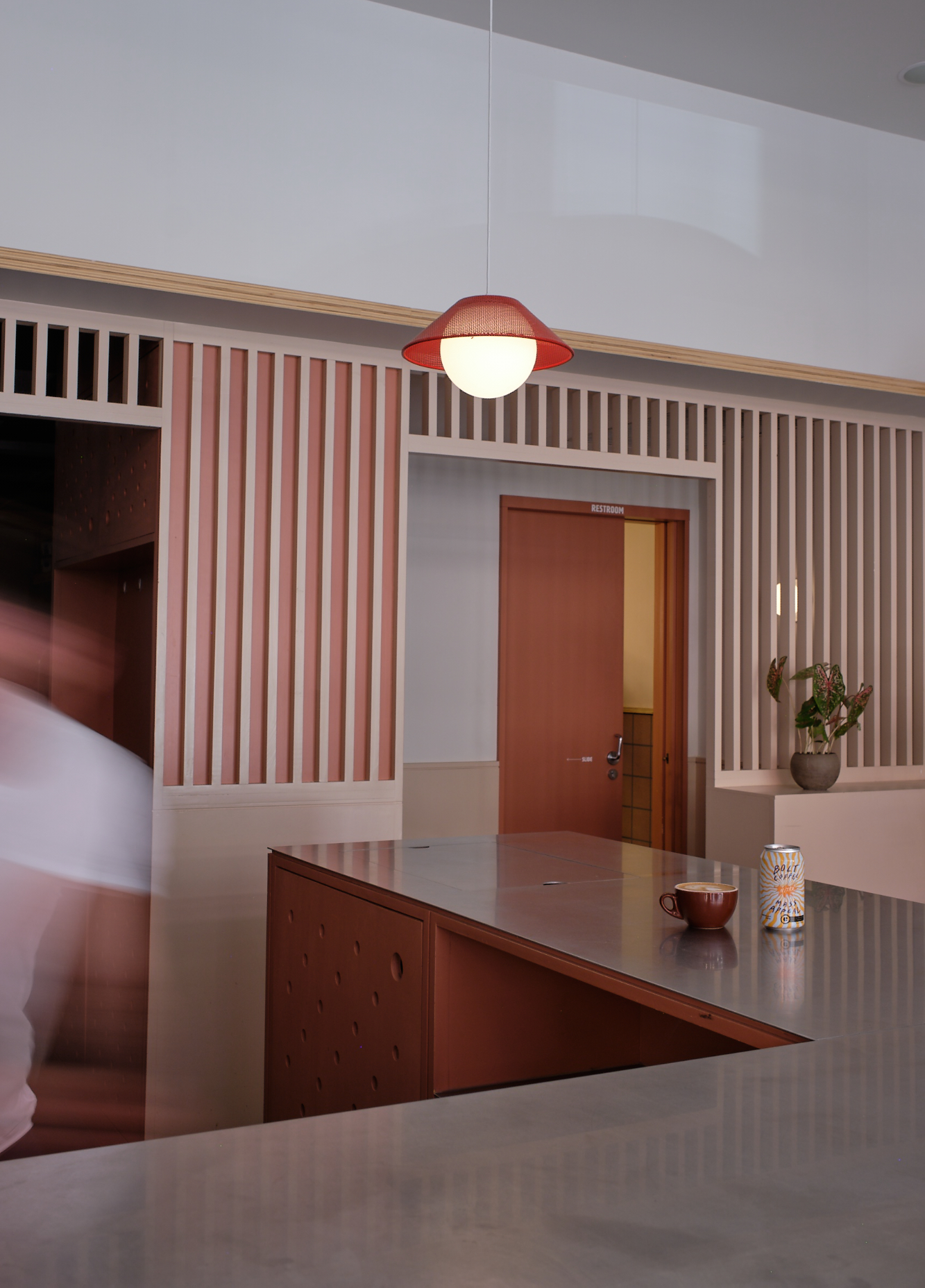 A coral-and-white slatted wall frames a doorway behind a café counter and red pendant light.