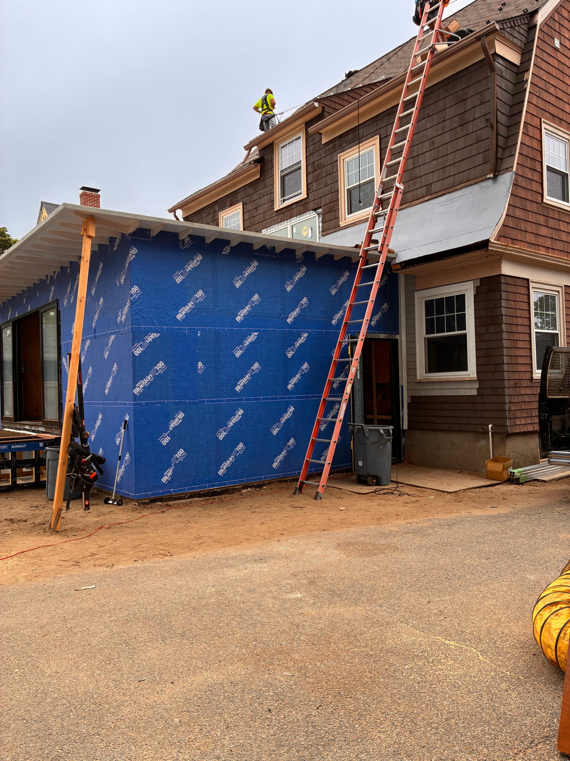 Addition under construction with blue wall sheathing, exposed roof edges and a ladder against the existing house.